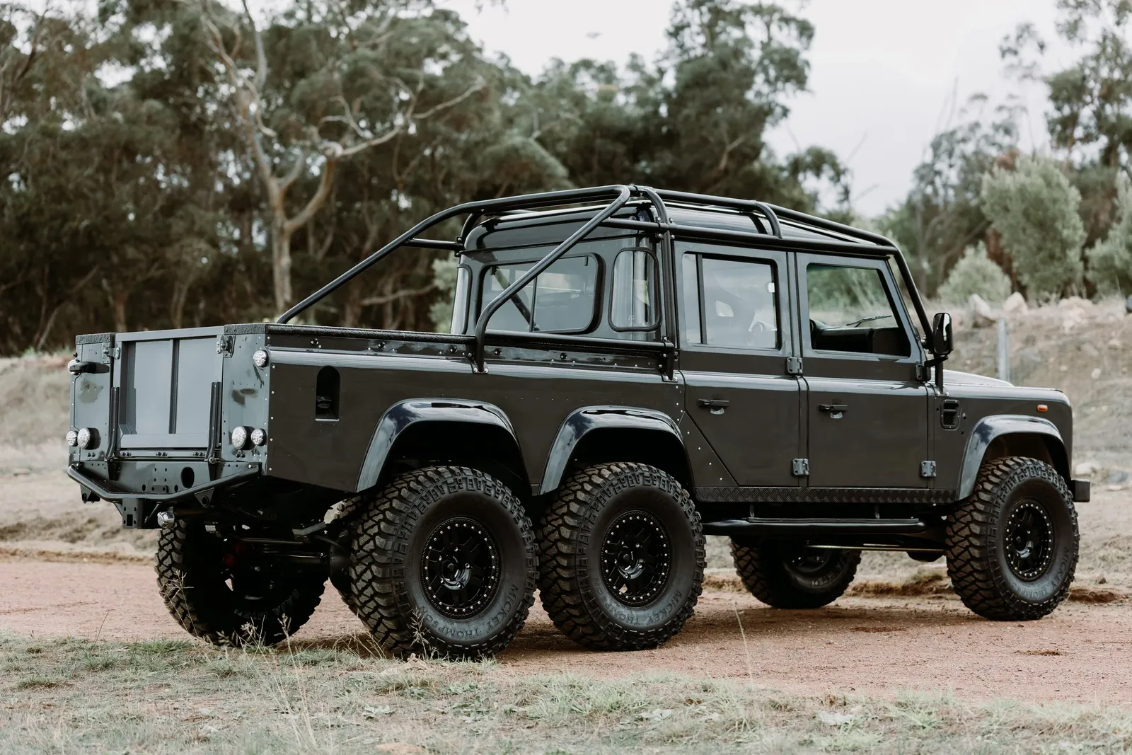 Dark grey six-wheel Land Rover Defender pickup with roll cage